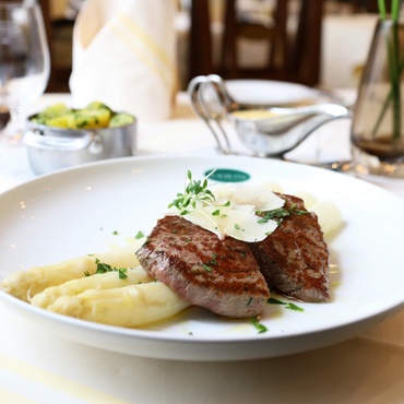 Gegrilltes Steak mit gehobeltem Käse, weißem Spargel und Kartoffelpüree auf einem weißen Teller in einer Restaurantumgebung.