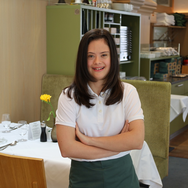 Junge Frau mit langen dunklen Haaren, die lächelnd und mit verschränkten Armen in einem Restaurant steht.