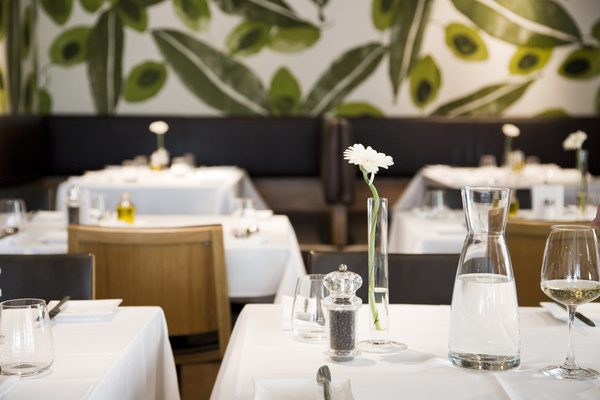 Modernes Restaurant mit weißen Tischtüchern, einfach gedeckten Tischen und einer begrünten Wandmalerei im Hintergrund.